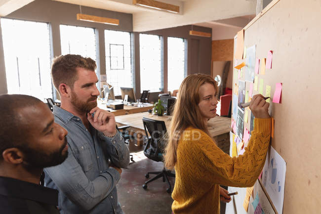 Side view close up of a young African American man and a young Caucasian man standing and watching their young Caucasian female colleague writing a note on an ideas board during a team brainstorm session at a creative office — Stock Photo