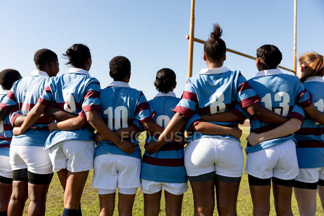 Rückansicht einer Mannschaft junger erwachsener multiethnischer Rugbyspielerinnen, die mit verbundenen Armen auf einem Rugbyfeld stehen und sich auf ein Rugbyspiel vorbereiten — Stockfoto