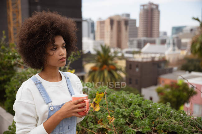 Вид збоку крупним планом молода змішана жінка, що стоїть на вулиці на балконі в місті, тримаючи чашку кави і дивлячись — стокове фото