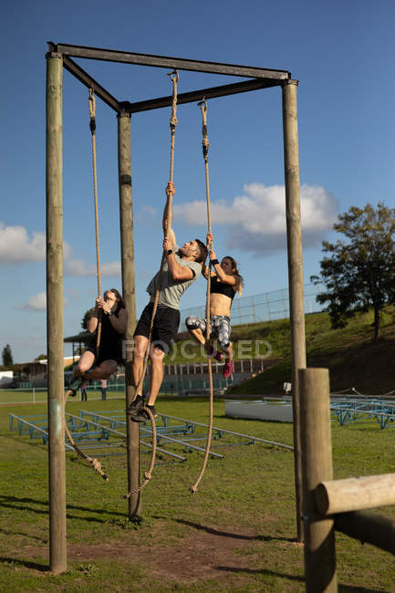 Vista frontal de dos mujeres caucásicas jóvenes y un hombre caucásico joven escalando cuerdas en un marco de escalada en un gimnasio al aire libre durante una sesión de entrenamiento de bootcamp - foto de stock