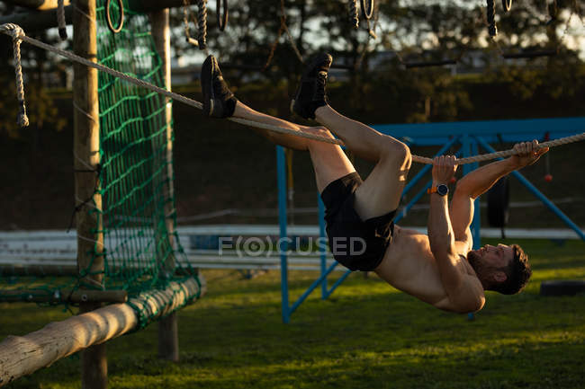 Vista lateral de um jovem caucasiano sem camisa pendurado em uma corda horizontal em uma armação de escalada em um ginásio ao ar livre durante uma sessão de treinamento de bootcamp — Fotografia de Stock