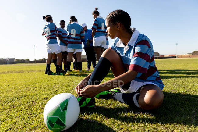 Vue de côté gros plan d'une jeune joueuse de rugby mixte adulte assise sur un terrain de rugby avec la balle, attachant sa botte et regardant ailleurs, avec ses coéquipières parlant ensemble en arrière-plan — Photo de stock