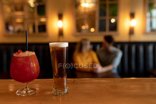 Vorderansicht eines glücklichen jungen kaukasischen Paares, das es sich im Urlaub in einer Bar gemütlich macht, sich umarmt und Händchen hält, im Vordergrund Bier und Cocktail — Stockfoto