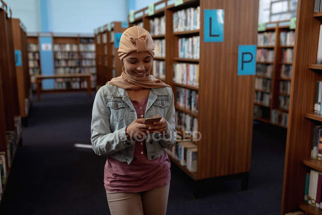 Передній вигляд біля молодої азійської студентки, одягненої в хіджаб, з допомогою смартфона в бібліотеці. — стокове фото