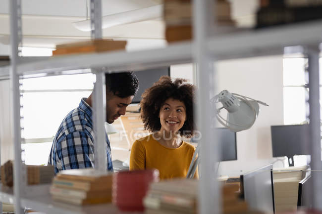 Vista laterale di un giovane uomo di razza mista e una giovane donna di razza mista seduta a una scrivania mentre discutono in un ufficio creativo — Foto stock