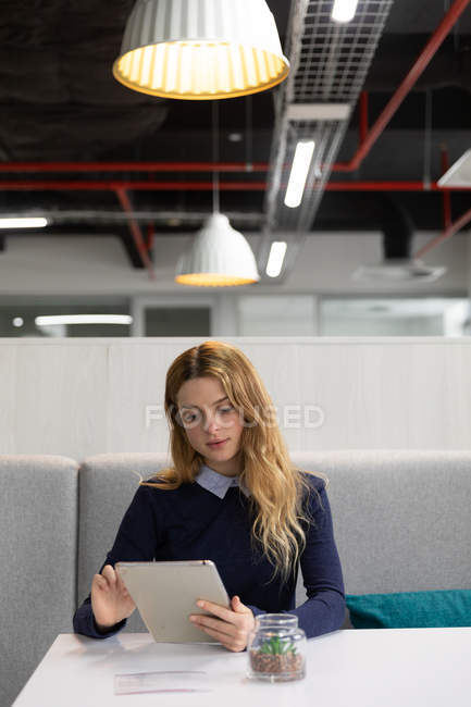 Vorderansicht einer jungen kaukasischen Frau, die mit einem Tablet-Computer an einem Tisch im Essbereich eines kreativen Unternehmens auf einer Bank sitzt — Stockfoto
