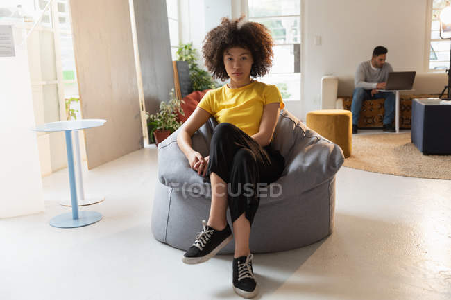 Porträt einer jungen Frau mit gemischter Rasse, die in die Kamera blickt, und eines jungen Mannes mit gemischter Rasse, der in einem Kreativbüro einen Laptop im Hintergrund benutzt — Stockfoto