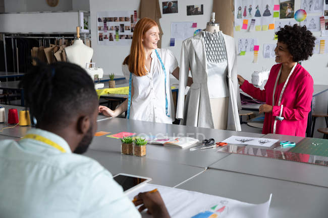 Rear view of a young African American male fashion student sitting and working on a design drawing while a young Caucasian female and a mixed race female fashion students work together with garments on a mannequin in a studio at fashion college — Stock Photo