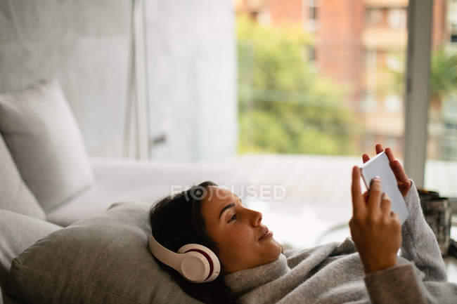 Вид збоку крупним планом молодий Кавказький жінка брюнетка лежачи на дивані носіння навушників і дивитися планшетний комп'ютер — стокове фото