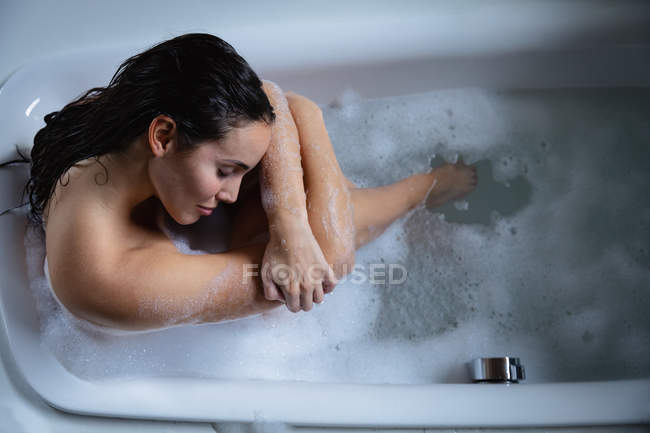 Sobre la cabeza de una joven morena caucásica sentada en un baño de espuma, sosteniendo sus piernas estiradas y apoyando su cabeza en sus brazos con los ojos cerrados - foto de stock