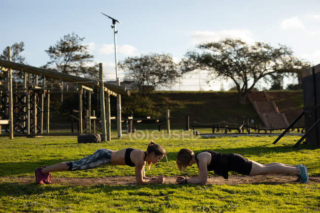 Vista lateral de dos jóvenes mujeres caucásicas haciendo el ejercicio de tablón en un gimnasio al aire libre durante una sesión de entrenamiento de bootcamp - foto de stock