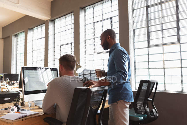 Vue latérale d'un jeune homme afro-américain tenant un café et un smartphone parlant avec un jeune collègue homme caucasien assis à un bureau avec son dos à la caméra à l'aide d'un ordinateur dans un bureau créatif — Photo de stock