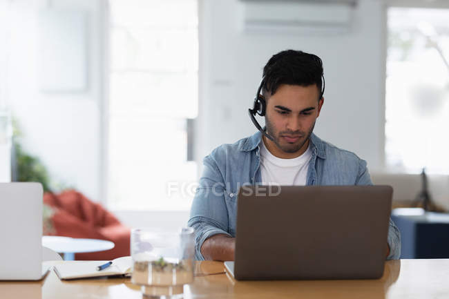 Передній вигляд молодого змішаного чоловіка, що сидить за столом у гарнітурі та використовує ноутбук у творчому офісі — стокове фото