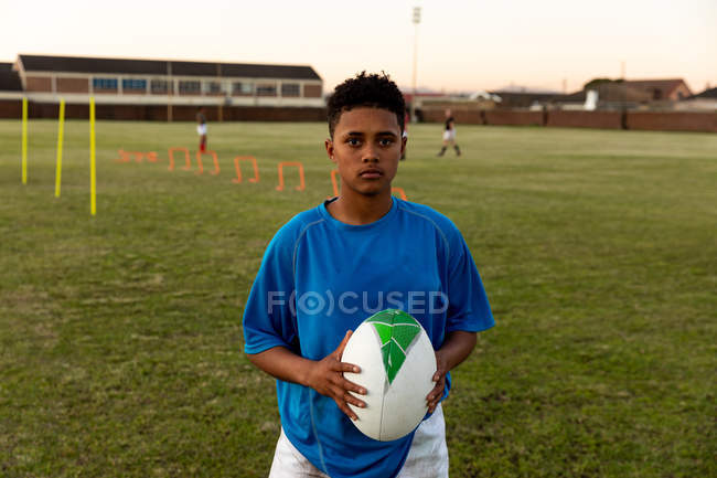 Retrato de una joven jugadora de rugby de raza mixta adulta parada en un campo de deportes sosteniendo una pelota de rugby durante una sesión de entrenamiento - foto de stock