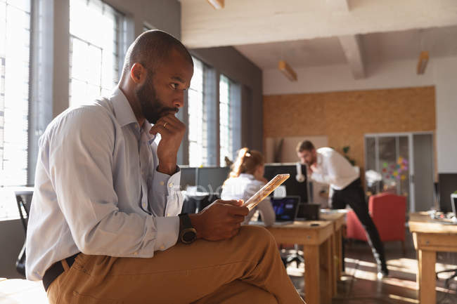Vista lateral de cerca de un joven afroamericano sentado en un escritorio usando una tableta y pensando en una oficina creativa, con dos colegas trabajando en segundo plano - foto de stock