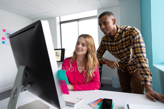Vista frontal de perto de um jovem afro-americano de pé e uma jovem mulher caucasiana sentada e conversando em uma mesa juntos olhando para um monitor de computador no escritório moderno de um negócio criativo — Fotografia de Stock