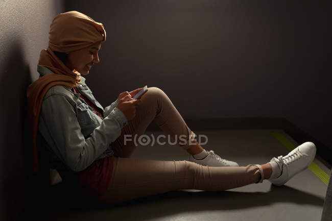 Vista lateral de cerca de una joven estudiante asiática usando un hijab usando un teléfono inteligente en una escalera - foto de stock