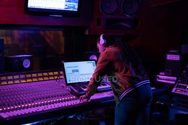 Вид збоку крупним планом молодий змішаної раси чоловічий звукорежисер, що стоїть за мікшерним столом у студії звукозапису, використовуючи комп'ютер та носячи навушники — стокове фото