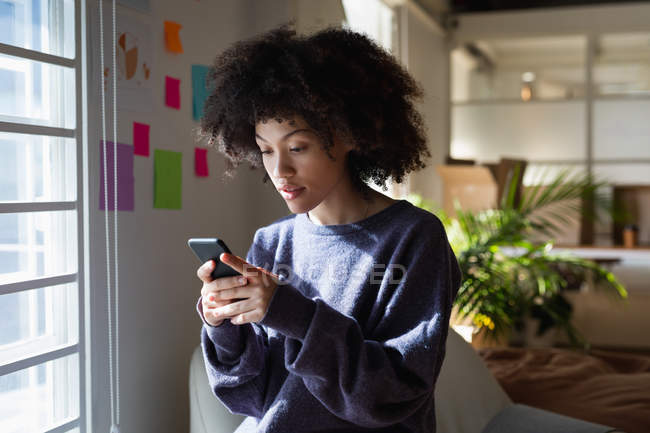 Seitenansicht einer jungen Frau mit gemischter Rasse, die auf einem Sessel sitzt und ein Smartphone in einem Kreativbüro benutzt — Stockfoto