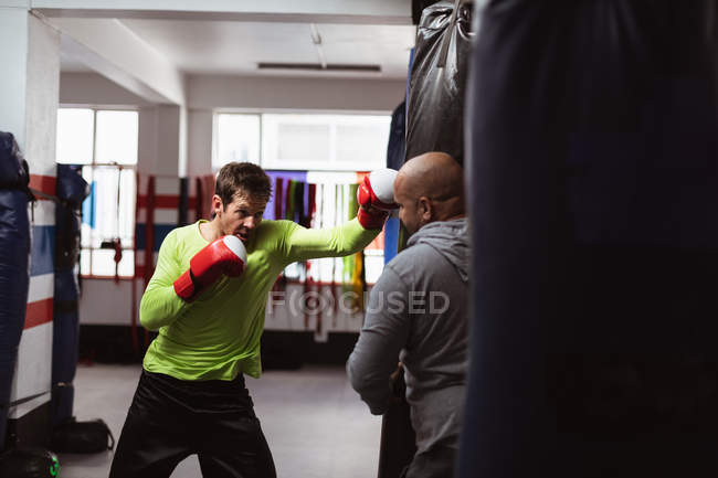 Vista frontal de um jovem boxeador caucasiano perfurando um saco de pancada segurado por um treinador masculino caucasiano de meia idade em um ginásio de boxe — Fotografia de Stock