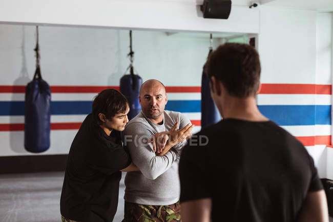 Передній вид закритий середнього віку Кавказький чоловічий інструктор, який проводить тренування з самооборони в боксерському залі, демонструючи підтримку молодого чоловіка змішаної раси, в той час як інший молодий чоловік дивиться на — стокове фото