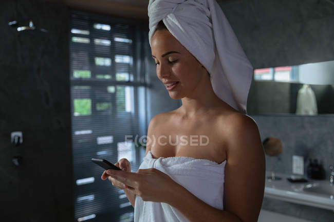Vista laterale di una giovane donna bruna caucasica che indossa un asciugamano da bagno e con i capelli avvolti in un asciugamano, usando uno smartphone in un bagno moderno — Foto stock