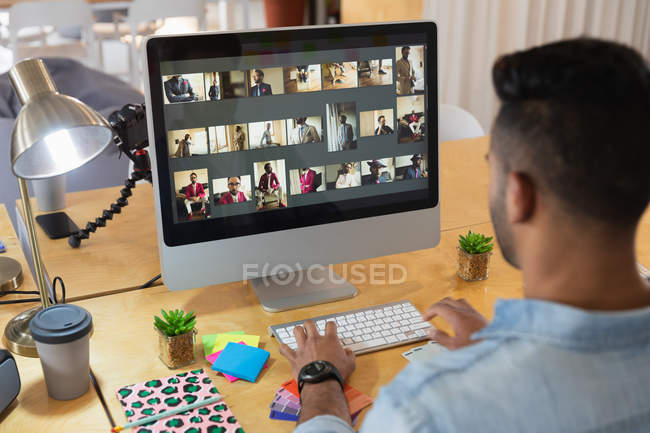 Vista trasera de un joven mestizo sentado en un escritorio y mirando una pantalla de computadora en una oficina creativa - foto de stock