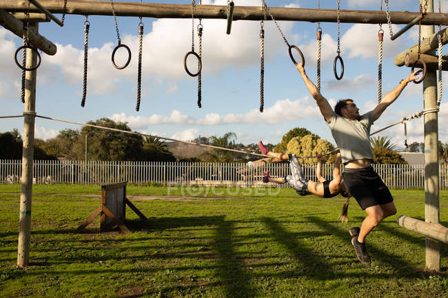 Vista frontal de um jovem caucasiano pendurado em anéis em uma estrutura de escalada em um ginásio ao ar livre durante uma sessão de treinamento de bootcamp — Fotografia de Stock