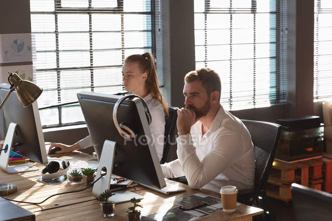 Side view of a young Caucasian woman and man sitting at a desk using computers in a creative office — Stock Photo