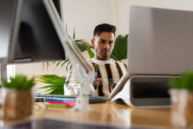 Вид спереду крупним планом молодий змішаний чоловік, що сидить за столом, використовуючи ноутбук у творчому офісі — стокове фото