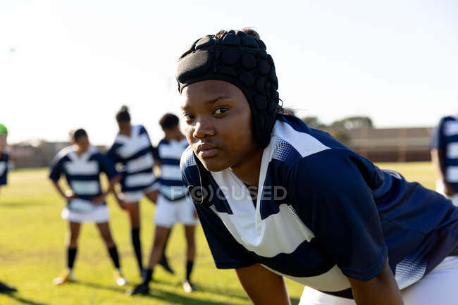 Vista frontal de uma jovem adulta de raça mista jogadora de rugby usando um protetor de cabeça em um campo de rugby inclinado para a frente e olhando para a câmera, com seus companheiros de equipe no fundo — Fotografia de Stock