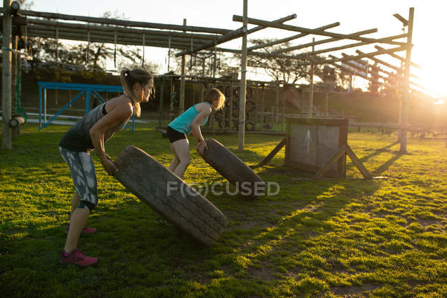 Vue latérale de deux jeunes femmes caucasiennes retournant des pneus dans une salle de gym extérieure lors d'une session d'entraînement bootcamp — Photo de stock