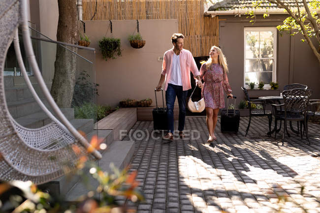 Vorderansicht eines glücklichen jungen kaukasischen Paares, das es sich im Urlaub gemütlich macht, mit Koffern vor einem Hotel stehend und Händchen haltend — Stockfoto