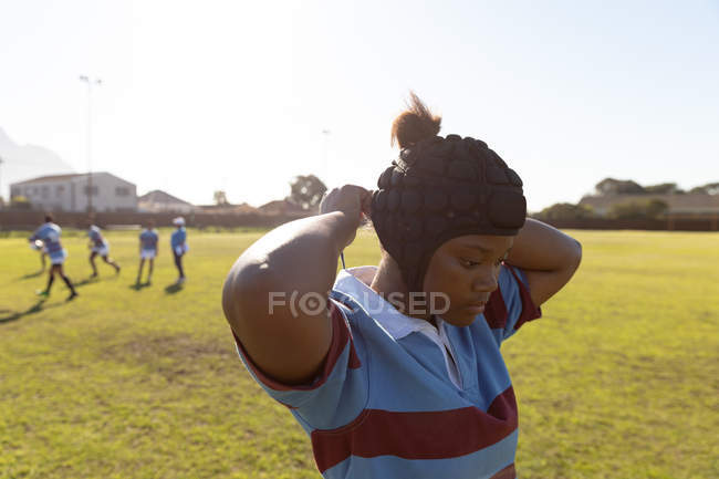 Vue de côté gros plan d'une jeune joueuse de rugby mixte adulte debout sur un terrain de rugby attachant son garde-tête, avec ses coéquipières en arrière-plan — Photo de stock