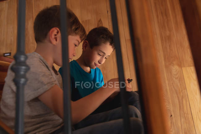 Вид сбоку на двух белых мальчиков, сидящих дома на лестнице, с помощью смартфона — стоковое фото