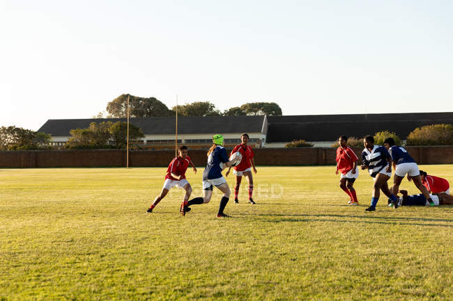 Vue latérale d'un groupe de jeunes joueuses de rugby multiethniques adultes courant sur le terrain pendant un match et poursuivant le joueur en possession du ballon — Photo de stock