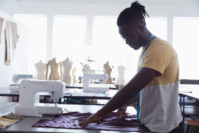 Vista lateral de un joven afroamericano estudiante de moda corte de tela mientras trabaja en un diseño en un estudio en la universidad de moda - foto de stock