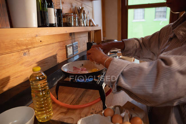Вид сбоку: молодая женщина смешанной расы разбивает яйцо в кастрюлю во время приготовления завтрака на кухне — стоковое фото