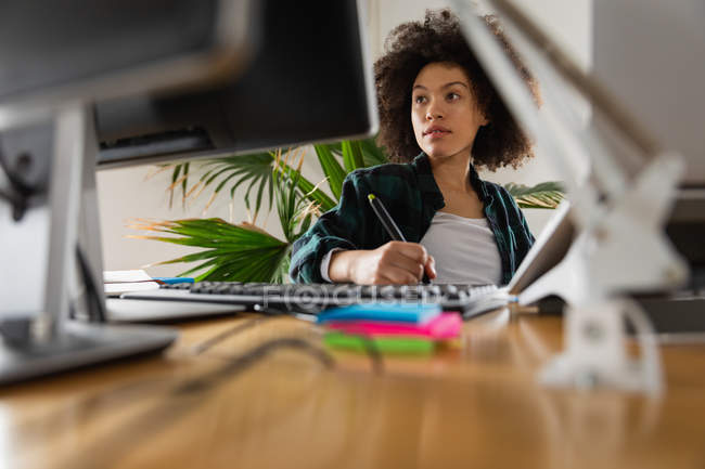 Nahaufnahme einer jungen Frau mit gemischter Rasse, die an einem Schreibtisch sitzt, auf einen Computermonitor blickt und in einem Kreativbüro einen Stift benutzt — Stockfoto