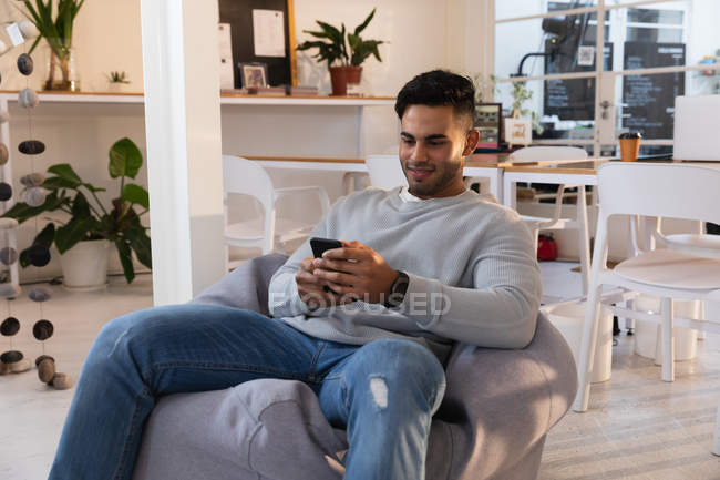 Вид спереду крупним планом молодий змішаний чоловік, що сидить на мішку з квасолею і використовує смартфон у творчому офісі — стокове фото