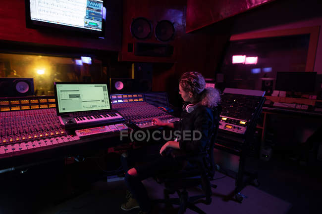 Вид сбоку на молодую кавказскую звукоинженершу, сидящую и работающую за микшерским столом в студии звукозаписи в окружении оборудования — стоковое фото