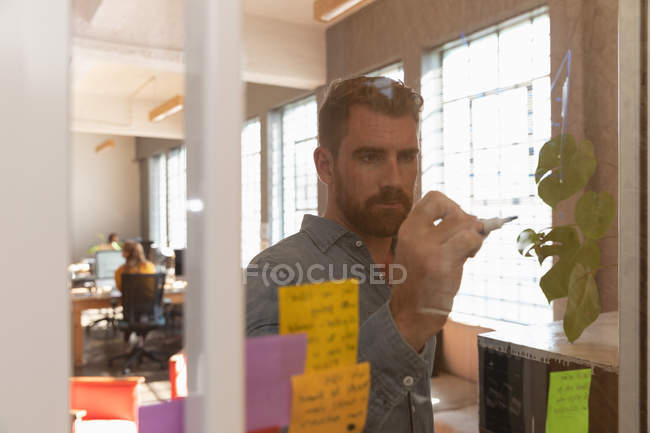 Вид спереди на молодого кавказца, пишущего заметки на стеклянной стене во время группового мозгового штурма в творческом офисе, через стеклянную стену, с коллегами, работающими на заднем плане — стоковое фото