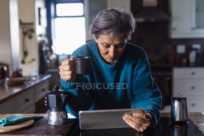 Vorderansicht einer älteren kaukasischen Frau in einer Küche, die Kaffee trinkt und einen Tablet-Computer mit Küchenschränken im Hintergrund benutzt — Stockfoto