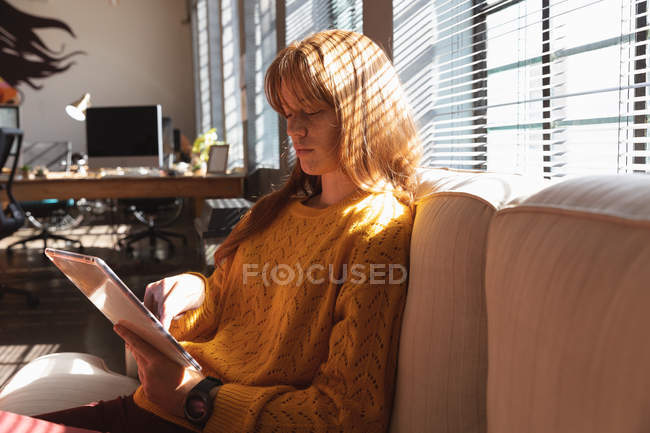 Vue de côté gros plan d'une jeune femme caucasienne assise sur un canapé à l'aide d'une tablette dans le salon d'un bureau créatif, rétroéclairée par la lumière du soleil — Photo de stock