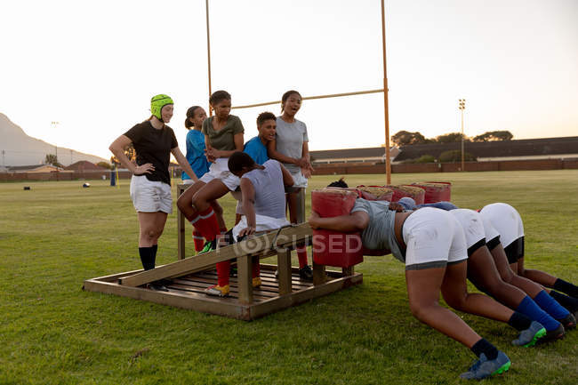 Seitenansicht einer Mannschaft junger erwachsener multiethnischer Rugbyspielerinnen auf einem Sportplatz bei einer Trainingseinheit, bei der drei der jungen Frauen mit einer Gedrängemaschine üben, während die anderen zusehen — Stockfoto