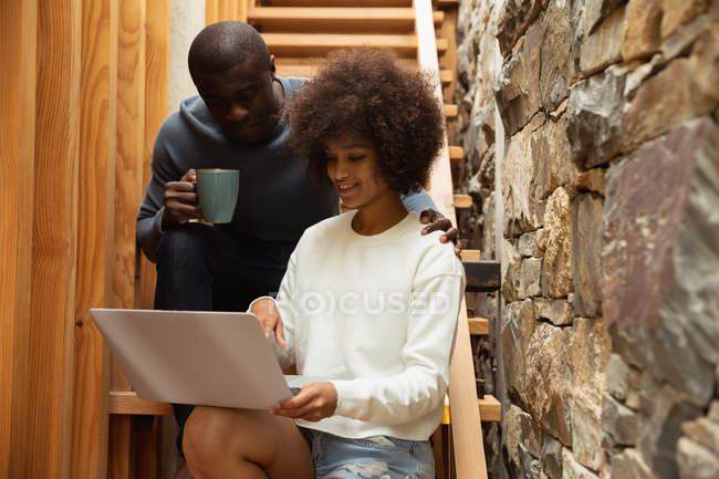 Vista frontale da vicino di una giovane donna di razza mista e un giovane uomo afroamericano che guarda un computer portatile e parla seduto sulle scale a casa . — Foto stock