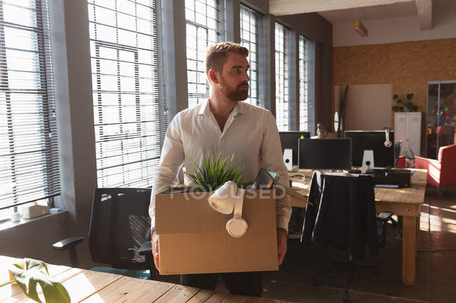 Передній погляд на молодого кавказького чоловіка, що стоїть за столом біля вікна у творчому кабінеті з картонною коробкою своїх речей, очистив свій стіл, щоб піти. — стокове фото