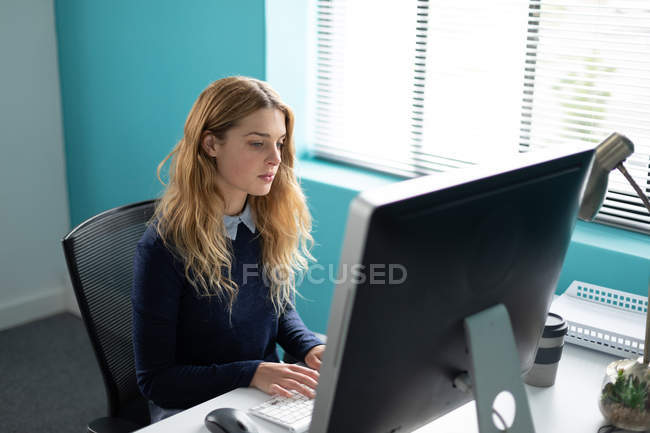 Vista lateral de una joven mujer caucásica sentada en un escritorio junto a una ventana usando una computadora, girando y sonriendo a la cámara en la oficina moderna de un negocio creativo - foto de stock