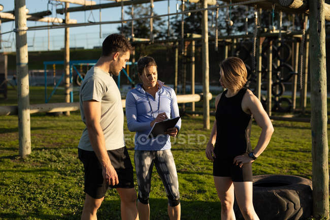 Front view of a young Caucasian woman and a young Caucasian man standing with a young Caucasian female instructor holding a clipboard during a bootcamp training session at an outdoor gym — Stock Photo