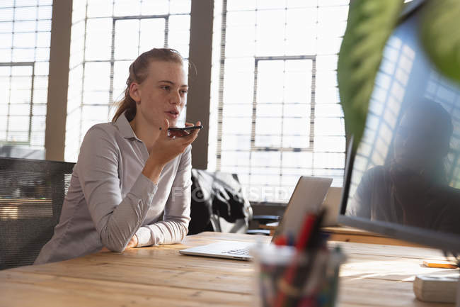 Вид сбоку. Молодая белая женщина сидит за столом за ноутбуком и разговаривает на смартфоне в креативном офисе. — стоковое фото